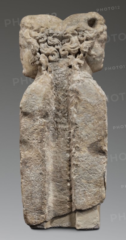 Double herm with heads of Apollo, unknown Roman workshop; herms; Apollo myth.; Roman sculpture, Antonine period 138–192; Roman Empire 27 BC–476; sculpture; height 67 cm, width 27 cm, depth 24 cm, weight 85.5 kg, The image shows a weathered stone sculpture of two human heads back-to-back atop a shared vertical block, with a rough textured surface and visible erosion. The heads have curly hair carved along the center where they meet, and both faces are shown in profile facing opposite directions. The stone displays a range of beige, tan, off-white, light brown, and gray tones, with darker gray shadows in crevices and lighter creamy areas on raised surfaces. The background is a smooth mid-gray.