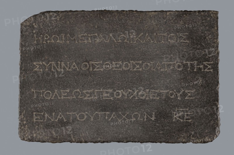 Greek dedicatory inscription, unknown author, tombstone inscription, 2nd half of the 2nd century BC, Egypt; Greek inscription; height 22.0 cm, width 32.5 cm, depth 7.0 cm, A dark rectangular stone slab with rough uneven edges resting against a plain neutral background, showing a textured surface with chiseled grooves forming horizontal rows; the slab is primarily dark gray with lighter gray scratches and worn areas, and the carved grooves appear as lighter gray lines that contrast with the darker surface, while the edges show some brownish and slightly lighter gray weathering.