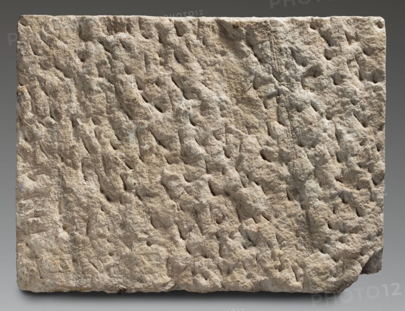 Tombstone of Marcus Julius Sistus with bilingual inscription, unknown Roman workshop; sepulchral funerary architecture, Greek inscriptions, Latin inscriptions, imperial freedmen. Rectangular marble slab with a very irregularly worked back surface. On the front surface are traces of numerous auxiliary lines vertical and horizontal, into which three lines of Latin text and three lines of Greek text are rather unevenly inscribed. Date: after 14 AD–end of 1st century 1st century. Place of origin: Italy, possibly Rome. Slab: height 42 cm, width 56 cm, depth 5 cm, weight 29 kg., A roughly rectangular block with an uneven, pitted surface showing a rough, weathered texture; the block displays a range of beige and tan tones with areas of light gray and small darker brown spots; the edges are irregular with one lower corner chipped, and the background is a smooth gradient of muted gray.