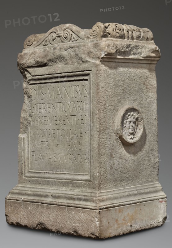 Funerary altar of Lucius Terentius Tarpius and Mucia Polla with Latin inscription, unknown Roman workshop; classified Gallic scenography; Medusa mythology; Latin inscriptions; sepulchral funerary architecture. At the top is a channel for a liquid offering. Sides decorated with representations of sacrificial vessels – a jug and a platter with the head of Medusa. End of the 1st century previously dated 2nd half of the 1st century–1st half of the 2nd century, Roman Empire 27 BC–476. Altar. Height 90 cm, width 59 cm, depth 44 cm, weight 480 kg., A stone rectangular monument with a weathered surface in shades of gray and beige, showing carved decorative scrollwork along the top edge and a raised framed panel on one side with an incised area. The front corner and edges appear chipped and rough, and the base is broader with layered molded profiles. One side features a small circular relief with a carved human face surrounded by stylized hair or foliage. The stone shows variations of light gra...