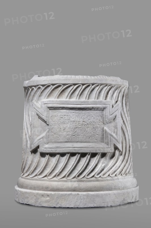 Urn of Gnaeus Pedanius Callistus with Latin inscription, unknown Roman workshop. A cylindrical funerary urn placed on a smooth plinth and a convex base. Its shape and decoration resemble a fragment of a column shaft, tapering towards the top. Entire surface decorated with S-shaped fluting strigillea, closed at top and bottom by a narrow frame. In the central part, slightly closer to the cornice than the base, is a tabula ansata with open handles containing the inscription. Inside the urn, a deep niche for ashes was chiseled out. The opening is topped by a molding decorated with radially arranged grooves; the place where the lid was attached is visible. Lid: missing; comparable examples have a conical or possibly domed roof. Inscription letter height 15 cm. Dating: beginning of the 3rd century previously dated late 2nd–3rd century AD. Probable findspot: Rome; place of origin: Roman Empire 27 BC–476. Urn Roman, weight 121.5 kg., A cylindrical stone object with carved spiral fluting aroun...