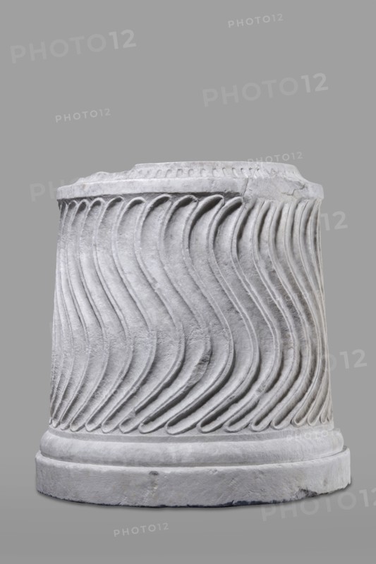 Urn of Gnaeus Pedanius Callistus with Latin inscription, unknown Roman workshop. A cylindrical urn placed on a smooth plinth and a convex base. Its shape and decoration resemble a fragment of a column shaft, tapering towards the top. Entire surface decorated with S-shaped fluting strigillea, closed at top and bottom by a narrow frame. In the central part, slightly closer to the cornice than the base, is a tabula ansata with open handles containing the inscription. Inside the urn a deep niche for ashes was carved out with a chisel. The opening is topped by a molding decorated with radially arranged grooves. The place where the lid was attached is visible. Lid: missing; a comparable urn has survived with its original conical lid, so it is assumed this urn likely had a similar conical or possibly domed lid. Inscription: letter height 15 cm. Dating: beginning of the 3rd century previously dated late 2nd–3rd century AD. Probable findplace: Rome; origin: Roman Empire 27 BC–476. Type: Roman u...