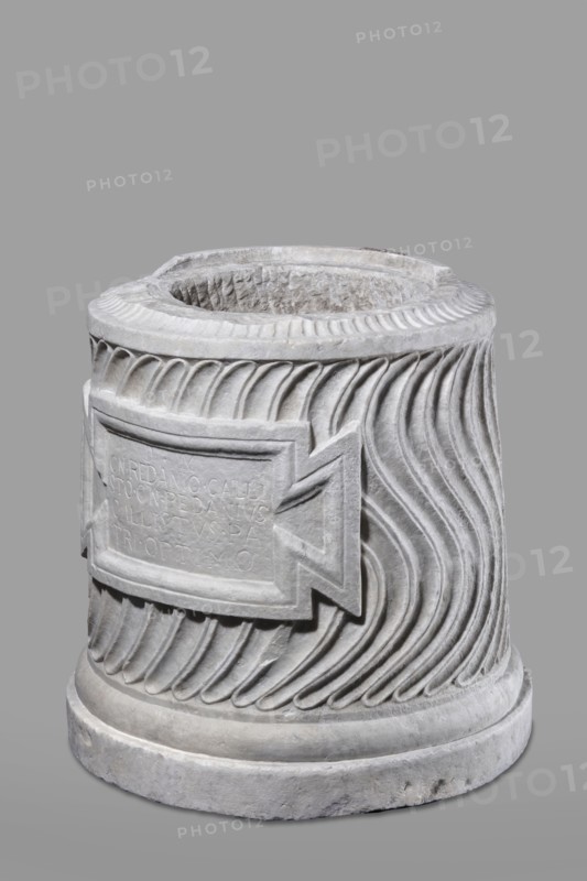 Urn of Gnaeus Pedanius Callistus with Latin inscription, unknown Roman workshop. A cylindrical urn placed on a smooth plinth and a convex base, its shape and decoration resembling a fragment of a column shaft, tapering towards the top. Entire surface decorated with S-shaped fluting strigillea, closed at top and bottom by a narrow frame. In the central part, slightly closer to the cornice than the base, is a tabula ansata with open handles containing the inscription. Inside the urn a deep niche for the ashes was chiseled out. The opening is topped by a molding decorated with radially arranged grooves; the place where the lid was attached is visible. Lid: missing; a comparable urn with its original conical lid suggests this urn likely had a similar conical or possibly domed lid. Inscription: letters height 15 cm. Dating: beginning of the 3rd century previously dated late 2nd–3rd century AD. Probable findspot: Rome; origin: Roman Empire 27 BC–476 AD. Urn Roman, weight 121.5 kg., A cylindr...
