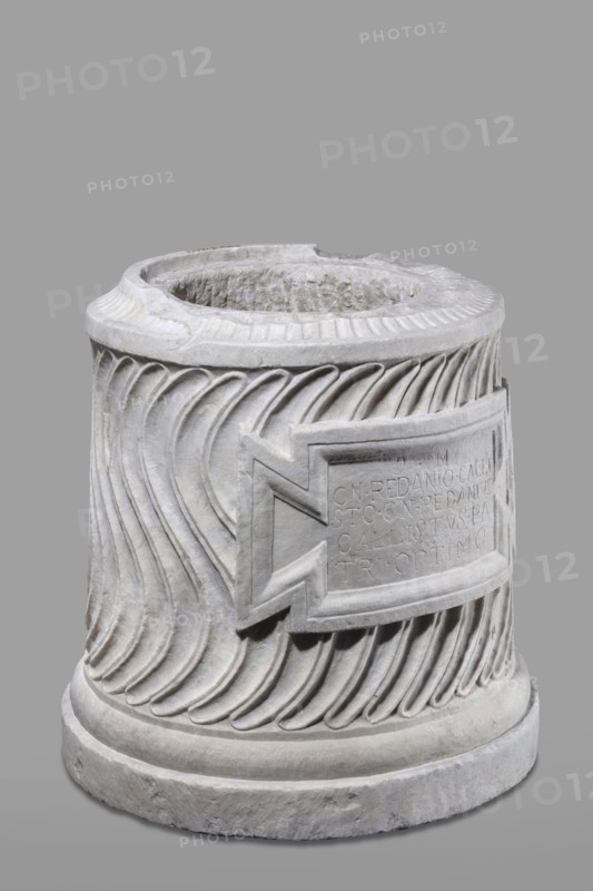 Urn of Gnaeus Pedanius Callistus with Latin inscription, unknown Roman workshop. A cylindrical urn placed on a smooth plinth and a convex base. Its shape and decoration resemble a fragment of a column shaft, tapering towards the top. Entire surface decorated with S-shaped fluting strigillea, closed at top and bottom by a narrow frame. In the central part, slightly closer to the cornice than the base, is a tabula ansata with open handles containing the inscription. Inside the urn a deep niche for ashes was carved out with a chisel. The opening is topped by a molding decorated with radially arranged grooves. The place where the lid was attached is visible. Lid: missing; comparable example with original conical lid suggests this urn likely had a similar conical or possibly domed lid. Inscription letters height 15 cm. Dating: beginning of the 3rd century previously late 2nd–3rd century AD. Probable findspot: Rome, Roman Empire 27 BC–476. Function: funerary urn. Weight 121.5 kg., A cylindri...