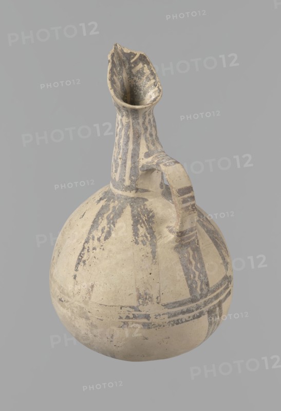 Jug, Excavated at Nea Paphos Cyprus. Cypriot pottery, Bronze Age White Painted IV, dated 1800–1650 BC Middle Cypriot period II–III. Cream clay with cream surface; geometric decoration in black. Pear-shaped belly, tall tapering neck, three-part spout, gently rounded base. Small vertical ribbon-like handle vise placed in an arc above the base of the neck; handle exterior decorated with three groups of three transverse lines each and painted sides; interior of spout similarly decorated. Neck covered with densely spaced straight and wavy lines, terminating at a horizontal line at the neck base. Upper body decorated with eight sets of two wavy lines radiating from the neck base, bordered by straight lines, extending to about one-third of the body height where they meet three horizontal lines at the vessel’s maximal bulge. Lower body shows four sets of three lines branching from these horizontals toward the base, irregularly spaced and angled. Good condition: two small rim fragments missing ...