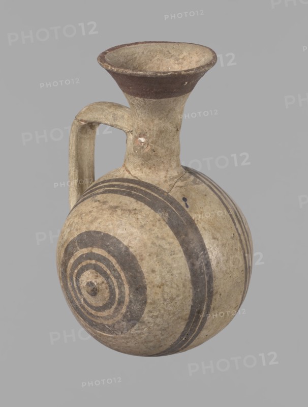 Cypriot gourd jug. A small vessel with a barrel-shaped gourd-shaped body, without a foot, with a spherical bottom. The center is marked with a small protruding point on both short sides. A single handle on the long side, mounted at the greatest bulge of the body and at the base of the neck, bent at a right angle. Quite long neck, strongly flaring, conical spout. Neck and handle slightly offset from the vessel axis; the handle is mounted at a slight angle, with a very clear trace of finger pressure on the right side where it is attached to the neck. Reddish-brown clay. Yellowish-brown engobe. Decoration in black-brown and matte red. The rim is decorated externally with a wide matte red band. The body is decorated on both sides with two groups of three concentric black-brown lines, of varying widths and spacing. In the groups on the shorter sides, the outer line is much wider than the others. Protruding points are marked in black-brown. Preservation condition: cracked and glued neck and ...