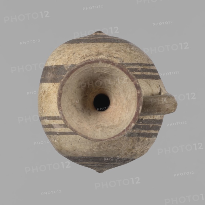 Cypriot gourd jug, A small vessel with a barrel-shaped gourd-shaped body, without a foot, with a spherical bottom. The center is marked with a small protruding point on both short sides. A single handle on the long side, mounted at the greatest bulge of the body and at the base of the neck, bent at a right angle. Quite long neck, strongly flaring, conical spout. Neck and handle slightly offset from the vessel axis; the handle is mounted at a slight angle, with a very clear trace of finger pressure on the right side where it is attached to the neck. Reddish-brown clay. Yellowish-brownish engobe. Decoration in black-brown and matte red. The rim is decorated externally with a wide matte red band. The body is decorated on both sides with two groups of three concentric black-brown lines, of varying widths and spacing. In the groups on the shorter sides, the outer line is much wider than the others. Protruding points are marked in black-brown. Preservation condition: cracked and glued neck a...
