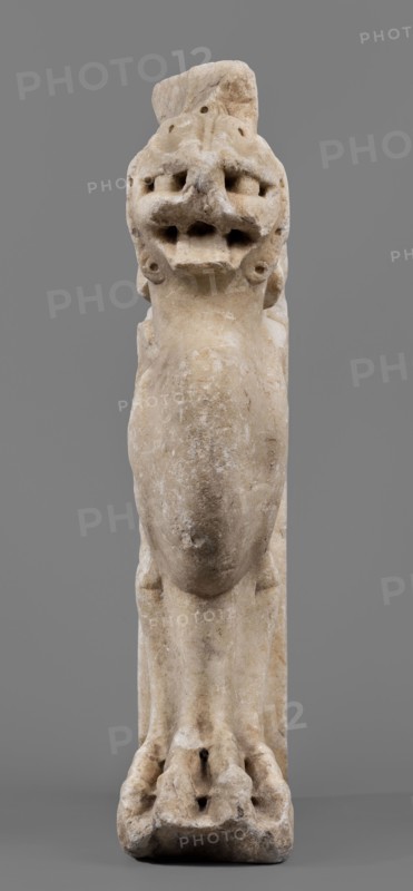 Bench support with a griffin figure, unknown Roman workshop; bench support decorated with a winged lion; mid-1st century, Roman Empire 27 BC–476; Roman sculpture; furniture; griffins myth.; table or bench support; height 46 cm, width 32 cm, depth 9 cm, weight 19.7 kg., A pale beige stone sculpture of an animal-like figure is shown against a solid gray background; the figure appears tall and vertical with a rounded elongated central body, a textured surface with weathered and slightly darker beige patches, and a head featuring deep-set hollowed areas and multiple rounded protrusions. The lower portion shows limb-like forms leading to a broad, irregular base with carved indentations and darker spots, and the overall colors visible are various shades of beige, off-white, and gray.