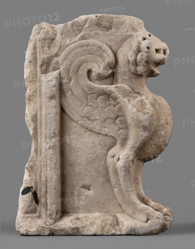 Bench support with a griffin figure, unknown Roman workshop, furniture, griffins myth., Roman sculpture, table or bench support decorated with a winged lion, ca. mid-1st century, Roman Empire 27 BC–476, furniture, sculpture; height 46 cm, width 32 cm, depth 9 cm, weight 19.7 kg, A carved stone relief shows a stylized winged creature in profile against a flat background. The creature has a rounded head with an open mouth and carved facial details, a curled decorative wing with scalloped feather patterns, a rounded body, and front legs ending in paws resting on a base. The stone surface displays weathering and rough texture with chips and worn edges. Visible colors include beige and light tan on the stone, small areas of darker gray and brown discoloration, a few spots of black, and a neutral gray backdrop.