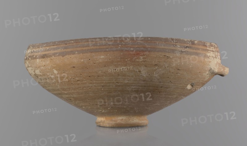 Bowl, excavations at Nea Paphos Cyprus — Bronze Age, 1650–1200 BC. Ceramic footed bowl/goblet on a low ring-shaped stem; walls diverge at a strongly obtuse angle and bend slightly inward at three-quarters height with a small horizontal vise at the bend. Light-sand clay with light-brown exterior paint and brown-red interior. Exterior painted decoration: narrow horizontal lines slightly darker than the ground, two horizontal purple stripes under the rim, a stripe covering the rim edge, and a belly stripe just below the rim. Interior with numerous light purple horizontal lines. Origin: Cyprus. Dimensions: height 8 cm, width 17 cm., A shallow, rounded ceramic bowl resting on a small foot, shown from the side against a neutral gray background, with a short protruding spout on one side; the bowl surface shows a weathered, rough texture with scratches and flaking, and has a base color of light brown and tan with areas of darker brown streaks and mottled white patches, with several horizontal ...