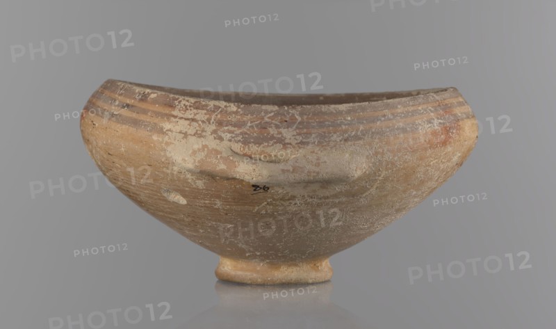 Bowl, excavations at Nea Paphos Cyprus - equivalent. Goblet on a low, ring-shaped stem; walls diverge at a strongly obtuse angle and bend slightly inward at three-quarters height with one small horizontal vise at the bend. Light-sand clay covered with light-brown paint outside and brown-red inside. Painted exterior decoration: narrow horizontal lines slightly darker than the ground, two horizontal purple stripes under the rim, a stripe covering the edge of the rim; belly just below the rim. Inside: numerous light purple horizontal lines. Cypriot pottery, Bronze Age, 1650–1200 BC. Vessel goblet, height 8 cm, width 17 cm., A shallow rounded bowl with a short foot, shown from the front against a neutral gray background, displaying an aged surface with worn patches and areas of flaking; the bowl is primarily tan and light brown with bands of darker brown near the rim, and there are patches of off-white residue and small speckles of darker brown and reddish-brown across its exterior.