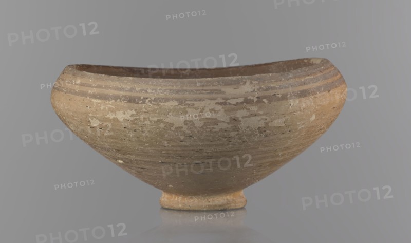 Bowl, excavations at Nea Paphos Cyprus; Bronze Age, 1650–1200 BC. Goblet on a low, ring-shaped stem; walls diverge at a strongly obtuse angle, bending slightly inward at three-quarters height with one small horizontal vise at the bend. Light-sand clay with light-brown paint outside and brown-red inside. Exterior painted decoration: narrow horizontal lines slightly darker than the ground, two horizontal purple stripes under the rim, a stripe covering the rim edge, and a belly stripe just below the rim. Interior has numerous light purple horizontal lines. Vessel type: footed bowl/goblet. Dimensions: height 8 cm, width 17 cm. Origin: Cyprus., A shallow rounded bowl with a small circular foot sits against a smooth gray background, the bowl showing a weathered surface with patches of lighter and darker areas; its overall color palette includes beige, tan, brown, cream, and hints of gray, with several horizontal bands near the rim and a worn, textured exterior.