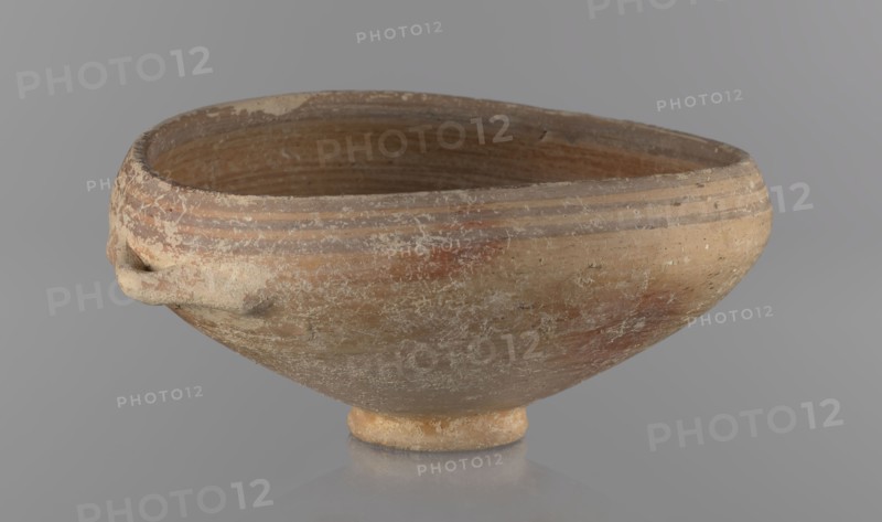 Bowl, excavations at Nea Paphos Cyprus - equivalent, vessels, Cypriot pottery, Bronze Age ceramics, footed bowl/goblet on a low ring-shaped stem; walls diverge at a strongly obtuse angle, bending slightly inward at three-quarters height with one small horizontal vise at the bend. Light-sand clay covered with light-brown paint on the outside and brown-red on the inside. Painted decoration on the outside: narrow horizontal lines slightly darker than the ground, two horizontal purple stripes under the rim; a stripe covering the edge of the rim; belly just below the rim. Inside: numerous light purple horizontal lines. 1650–1200 BC, Cyprus, vessel goblet, height 8 cm, width 17 cm, A shallow rounded bowl with a small foot, showing a weathered surface and textured patches, with a small applied handle-like protrusion on one side; the interior and exterior surfaces display concentric ridges near the rim and worn horizontal striations. The visible colors include shades of beige, tan, light brown...