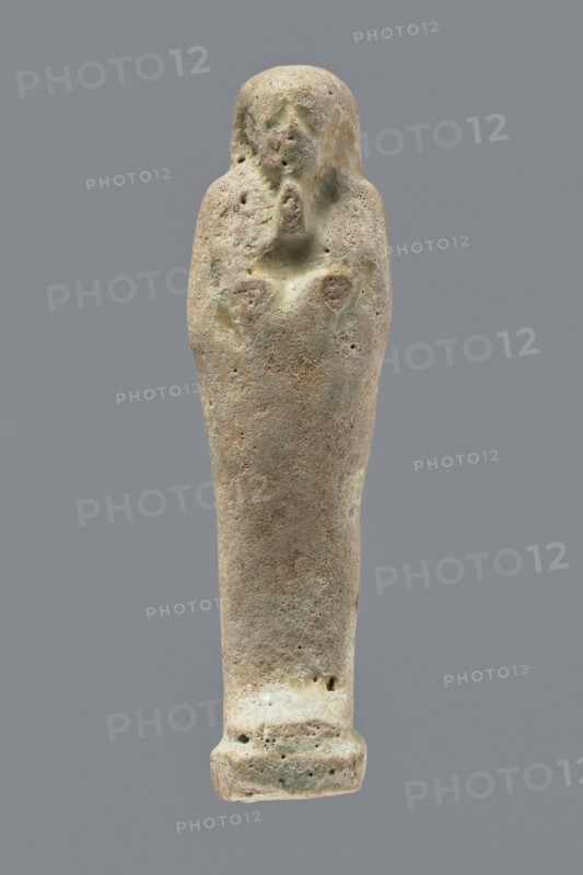 Ushabti, mummy-shaped figurine, funerary gift, hoes. Mummy-shaped figurine standing on a base, back supported by a narrow flat pillar. Arms crossed on the chest, right arm folded over the left; both hands hold hoes. Smooth tripartite wig, round face, facial details illegible, long straight beard. Not inscribed. Egypt; ushabti; height 11.8 cm, width 3.3 cm., A single elongated statue-like object stands against a plain gray background, with a rounded top tapering down to a flared base; the surface appears rough and pitted with a weathered texture and irregular small holes. The object shows a range of muted colors including beige, light brown, pale green, and hints of gray, with darker brown spots and lighter creamy areas. Near the upper portion there are shallow, worn contours suggesting a simplified facial area and crossed arms, with subtle raised and recessed areas around the midsection. The overall shape is narrow and slightly tapered, and the base is broader and slightly squared with...