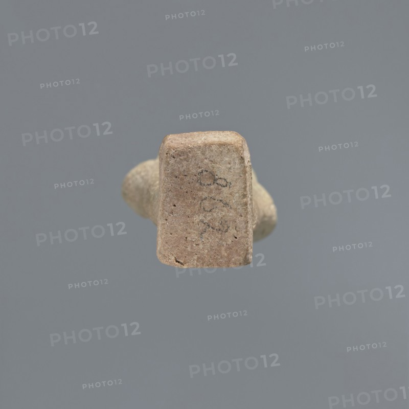 Ushabti, Mummy-shaped figurine, standing on a base, the back supported by a flat pillar. The arms are crossed on the chest and placed at the same level, some distance apart. In each hand are hoes; in the right hand is also a string from which a basket is slung over the left shoulder. The tools are engraved. On the head is a smooth, three-part wig. The face is elongated, with clearly defined eyes, a long nose, and a slight smile on the mouth. A long, straight beard. Not inscribed. 525–343 BC; Late Period, 27th–30th Dynasty, Egypt. Ushabti, height 9.6 cm., A small, irregularly shaped object with a rough, sandy surface in tan and light brown tones is centered against a uniform gray background; the object shows a rectangular face facing forward with some darker speckles and faint dark markings, and a rounded, tapering body extending behind it in similar tan and beige colors.