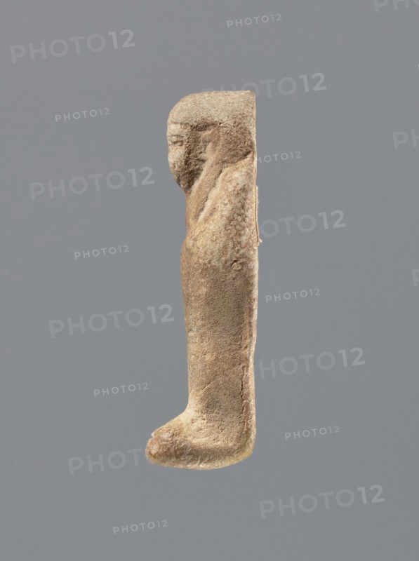 Shabti, Mummy-shaped figurine, standing on a base, the back supported by a flat pillar. Arms crossed on the chest, holding hoes in both hands. In the right hand a string from which a basket is slung over the left shoulder. Slightly convex tools. Smooth, tripartite wig. Round face with clearly defined large eyes; mouth and nose not visible. Not inscribed. 525–343 BC; Late Period, 27th–30th Dynasty, Egypt. Shabti; height 7 cm., A worn carved figure shown in profile with a rounded head and a simplified facial profile, a sloped shoulder and an elongated, straight torso that tapers down to a rounded foot-like base; the surface appears textured and pitted. The visible colors are shades of brown, tan, beige, and hints of pale yellow, set against a neutral gray background.