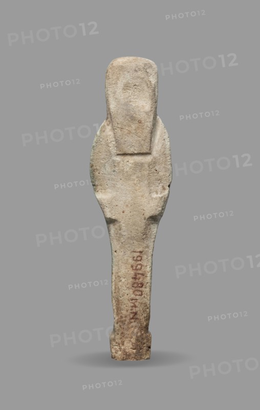 Ushabti, Mummy-shaped funerary figurine standing on a base, the back supported by a flat pillar. Arms crossed on the chest with hands visible from under the shroud; right hand slightly above the left. Left hand holds a pickaxe, right hand holds a hoe and a string from which a basket is slung over the left shoulder. Convex tools. Smooth three-part wig; triangular face with clearly marked large eyes, wide nose, mouth in a slight grimace. Not inscribed. Late Period, 664–332 BC, Egypt. Shabti; height 9.3 cm, width 2.5 cm., A vertical, elongated stone object with a rounded top and tapered lower section, shown against a plain gray background. The surface of the object is rough and weathered, displaying mostly beige and light tan tones with small patches of darker brown and faint greenish discoloration. The silhouette suggests a slightly wider midsection with subtle indentations along the sides and a flattened upper area. There are areas of reddish-brown staining on the lower portion and some...