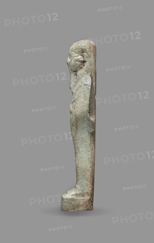 Ushabti, Back pillars, wigs, funerary figurines, hoes, grave goods, mummy-shaped figurines, standing on a base, the back supported by a flat pillar. Arms crossed on the chest. Hands visible from under the shroud, the right slightly above the left. In the left hand a pickaxe, in the right a hoe. In the right hand also a string, on which a basket is slung over the left shoulder. Convex tools. On the head is a smooth, three-part wig. Triangular face, clearly marked large eyes, a wide nose, mouth in a slight grimace. Not inscribed. 664–332 BC; Late Period, Egypt. Shabti; height 9.3 cm, width 2.5 cm., A narrow vertical statuette of a standing human figure shown in profile against a plain background, with a smooth elongated shape and simplified features including a defined head, shoulders, and torso; the surface appears textured and worn with areas of roughness and small pitting, and the form narrows toward the feet which rest on a small rectangular base; visible colors include muted green, ...