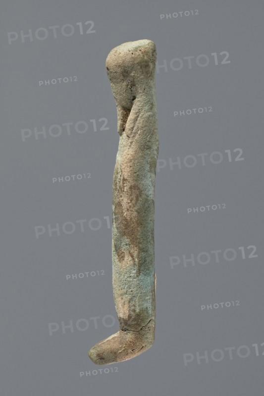 Ushabti, Mummy-shaped figurine, very schematically shaped. Hands crossed on the chest. On the head is a long, tripartite wig. Facial details illegible. Not inscribed. ca. 1069–ca. 656 BC; Third Intermediate Period, Egypt. Ushabti; height 9.4 cm., A single elongated vertical object with an irregular rounded top and a curved hooked end, showing a rough, pitted surface texture and small surface indentations; the object displays a mix of green, bluish, brown, beige, and gray tones across its surface, with patches of darker brown and lighter beige creating a mottled appearance, and it is set against a uniform gray background.