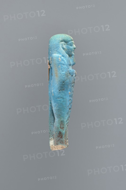 Ushabti, Mummy-shaped figurine, back supported on a flat pillar. Feet missing. Arms crossed on the chest, right folded over left. Hoes in both hands. In the right hand, a string on which a basket is slung over the left shoulder. Slightly convex tools. On the head, a smooth, three-part wig. Round face, clearly marked large eyes, wide nose, mouth in a slight grimace. Engraved inscription, placed in one column on the front of the legs; text illegible. 404–343 BC; Late Period, 28th–30th Dynasty, Egypt. Ushabti; height 7 cm., A small sculpted figure viewed in profile against a plain gray background, predominantly turquoise blue with areas of lighter blue and pale green patina, showing a human form with a rounded head, facial features, and a long body wrapped or clothed; there are a few rougher, weathered spots of brown and beige, and a small patch of white near the lower end, with some subtle darker blue and gray speckling across the surface.
