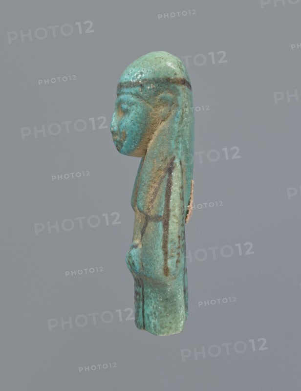 Ushabti, mummy-shaped figurine, preserved upper fragment from the waist up. Arms crossed on the chest, hands placed at the same level, next to each other, holding hoes. Rectangular basket on the back. Long, smooth, three-part wig with a headband; triangular face with proportional details; eyes slightly closed. Headband and facial details painted. Painted inscription in a column separated by vertical lines on the front of the legs, starting just below the folded hands; text illegible. Shabti, c. 945–c. 715 BC, Third Intermediate Period, 22nd–23rd Dynasty, Egypt. Height 5.8 cm., A small sculpted figure shown in profile against a plain gray background, predominantly turquoise with areas of greenish-blue and mottled brown, depicting a human-like head with a smooth rounded top and long hair or headdress that extends down the back; the face has a pronounced nose and chin, a slightly rounded cheek, and a subtle ear shape, with the body beneath showing a narrow torso and arms positioned along ...