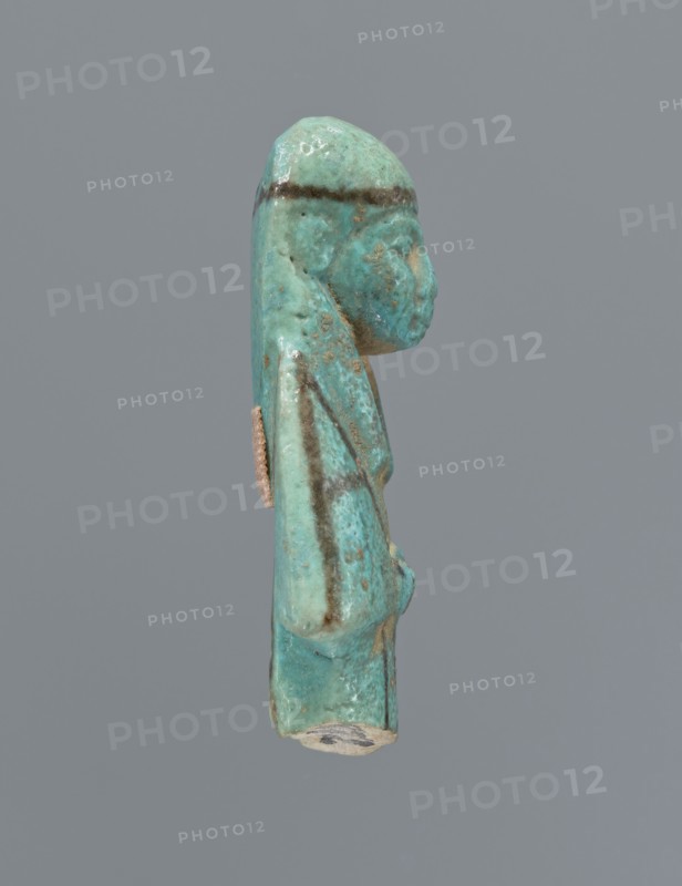 Ushabti, mummy-shaped figurine, preserved upper fragment from the waist up. Arms crossed on the chest, hands placed at the same level, next to each other, holding hoes in both hands. Rectangular basket on the back. Head wears a long, smooth, three-part wig with a headband. Triangular face with proportional details; eyes slightly closed. Headband and facial details painted. Painted inscription placed in a column separated by vertical lines on the front of the legs, starting just below the folded hands; text illegible. c. 945–c. 715 BC, Third Intermediate Period, 22nd–23rd Dynasty, Egypt. Height 5.8 cm., A small turquoise-green figurine viewed in profile against a plain gray background, showing a human-like head with a rounded top and a pronounced nose, a long headdress or hair that extends down the back, and a slightly protruding torso with an arm folded against the body; the surface shows areas of darker green and brownish streaks and spots, along with patches of lighter green and some...