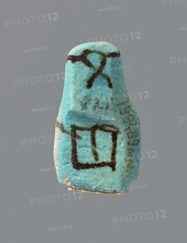 Ushabti, mummy-shaped figurine, preserved head and fragment of torso. Arms crossed on the chest, hands placed at the same level, next to each other, holding hoes in each hand. On the back, a square basket is slung on strings over both shoulders. On the head is a long, smooth, tripartite wig with a headband. Triangular face, details almost illegible. Headband and tools painted. ca. 945–ca. 715 BC; Third Intermediate Period, 22nd–23rd Dynasty, Egypt; height 3.7 cm, A small object with a turquoise blue surface suspended against a gray background, showing a few dark brown markings on its upper and middle areas and a darker brown outline forming a rough rectangular shape on its lower half, with some pale beige material visible along its bottom edge and a few tiny darker speckles across its surface.
