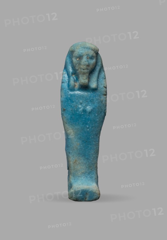 Ushabti, Wigs, funerary figurine, grave goods, mummy-shaped figurine. The arms are not distinct, with a slight convexity of the clenched hands. Delicate outline of tools on the arms. On the head is a smooth, tripartite wig. The face is large, round, with large eyes, a wide nose, and a slightly grimacing mouth. Not inscribed. Date: 525–332 BC, Late Period, 27th–30th Dynasty, Egypt. Ushabti; height 5.1 cm, width 1.5 cm., A small statuette shaped like a wrapped figure stands against a plain background, predominantly blue with areas of lighter and darker blue and small patches of brownish discoloration; the figure has a rounded head with simple facial features and a headdress-like form, a tapered body resembling a shroud, and a slightly wider base, with a rough textured surface and a few small pits and irregularities.