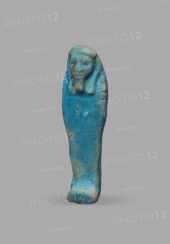 Ushabti, Wigs, funerary figurine, mummy-shaped figurine. The arms are not distinct, with a slight convexity of the clenched hands. Delicate outline of tools on the arms. On the head is a smooth, tripartite wig. The face is large, round, with large eyes, a wide nose, and a slightly grimacing mouth. Not inscribed. 525–332 BC; Late Period, 27th–30th Dynasty, Egypt. Ushabti; height 5.1 cm, width 1.5 cm., A small sculpted figure shown against a plain gray background, rendered in bright turquoise blue with areas of lighter blue and some pale beige discoloration; the figure has a rounded head with a suggestion of facial features and a headdress-like form extending down the sides, a narrow elongated body tapering toward a slightly thicker base, a textured surface with small pits and worn spots, and soft shadow beneath it.
