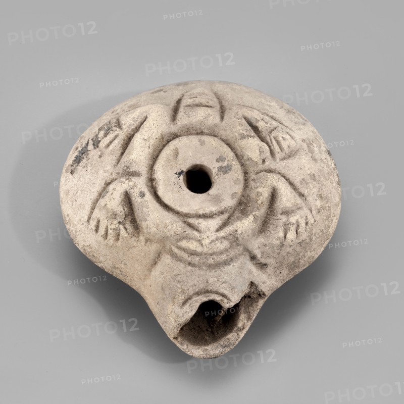 Lamp, Oil lamp with a circular container and a slightly forward burner. Decorated on the top with a schematic image of a frog surrounding the filler hole. The letter A is carved on the bottom. 3rd–4th century, Egypt. Oil lamp; height 3.4 cm, width 8.1 cm, depth 7.3 cm., A round, worn object with a central circular hole and a broken, curved opening at one edge, made of a light gray-beige material with darker gray spots and patches; its surface shows carved, raised decorative motifs forming curved lines and triangular shapes around the center, and the object casts a soft shadow on a smooth light gray background.
