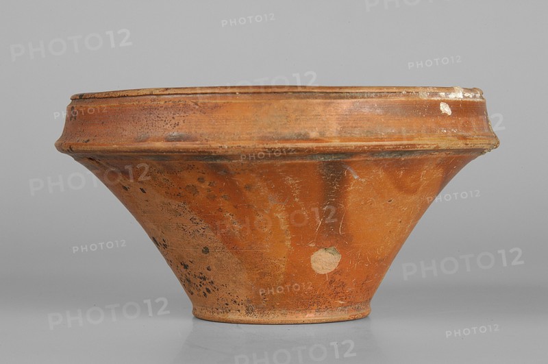 Bowl, Ancient Black Sea region, end of 1st–beginning of 2nd century AD, Crimea, vessel, height 6.1 cm, width 11.2 cm, A low, wide shallow bowl with a flared profile sits on a smooth surface; the vessel shows an earthy, terracotta orange color with darker brown and black spotting and areas of wear, along with a few paler, almost beige patches where the surface is abraded. The rim is slightly raised and has a similar orange-brown tone with small chips and rough edges. The body of the bowl has a matte, textured finish with scratches and irregular discoloration, and the base is narrower and slightly darker than the upper portion. The background is a uniform light gray.
