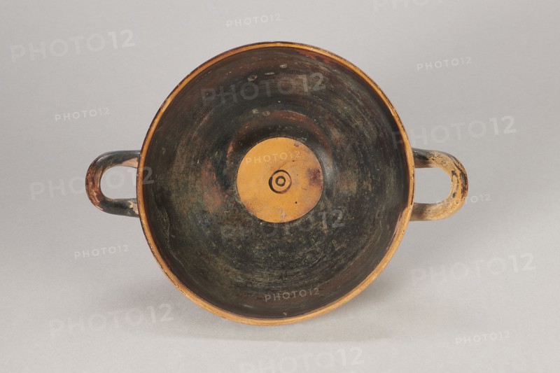 Black-figure kylix with Greek inscription, Archaic kylix form between a band/ribbon kylix and an eye kylix, 540–520 BC, Attica, kylix vessel, height 16.2 cm, width 22.2 cm, A top-down view of a shallow round bowl with two loop handles on opposite sides, showing interior surfaces that are predominantly dark with worn areas revealing brownish and ochre tones, a central circular ochre area with concentric rings, handles that are mottled with dark brown, black, and ochre, and a neutral light gray background.