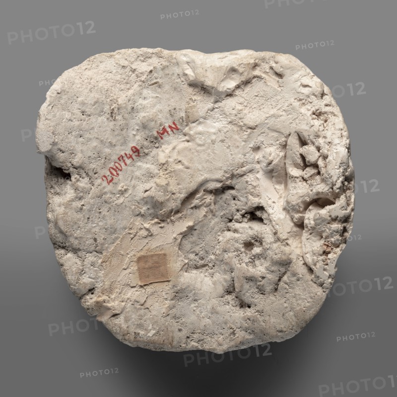 Mummy mask, Round face modeled in stucco with large eyes, a wide nose, a mouth in a slight half-smile, no ears. Face height: 12 cm. Sarcophagus mask, height 12 cm, width 11.7 cm, weight 0.342 kg. 2nd century, Roman period, Egypt. Materials: stucco. Function: grave good; Egyptian art, masks, faces., A roughly circular, irregularly shaped pale object with a rough, textured surface showing areas of coarse and smoother plaster-like material; the surface has a variety of depressions, ridges, and small cavities and appears worn and crumbly in places; a small square patch of a slightly darker beige fabric-like material is affixed to one area; there are traces of a red pigment on the surface but no readable markings are described; the overall colors visible are shades of off-white, cream, beige, light gray, and touches of darker gray against a neutral medium-gray background.