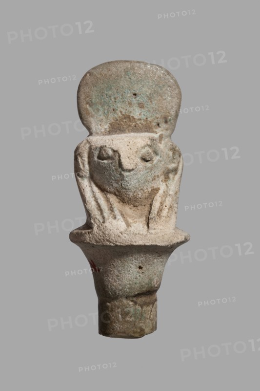 Amulet - falcon head on a column; papyrus capital of the column shows a falcon head in a striped wig topped with a disc, with a pendant at the back. 332–30 BC, Ptolemaic Period, Egypt. Amulet; height 3.6 cm, width 1.5 cm., A small sculpted object resembling a stylized crouching figure with a rounded top, rough textured surface, and simplified facial features; the piece shows a head with a flat forehead area above recessed eyes and a suggestion of limbs folded close to the body, all carved in a single piece that tapers into a cylindrical base. The colors visible are pale beige, light tan, muted green, and small areas of brown and gray, with subtle mottling and weathered patches across the surface.