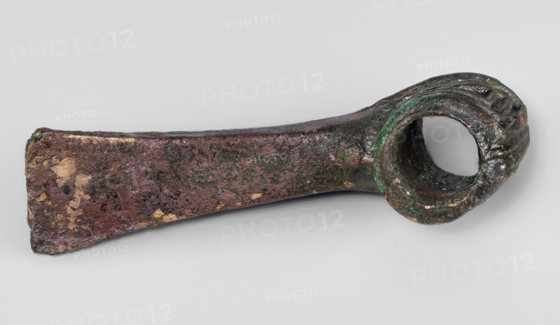 Pounder, unknown Near Eastern workshop; pounder with a low, ribbed, cylindrical socket and a blade in the form of a massive wedge set at a slight angle to the shaft. 1st half of the 2nd millennium BC; Middle Bronze Period. Western Iran find/origin. Material: metal; function: weapon. Dimensions: height 4.6 cm, width 12.5 cm, depth 3.2 cm; weight 324 g. Geometric ornaments; edged weapons; Eastern Gaulish scenography., An aged metal object with a tapered rectangular shaft leading to a rounded hollow loop; the surface shows rough, pitted texture with patches of reddish-brown, dark brown, green, and some areas of lighter tan or gold tones, and the loop end has visible striations and interior darkening.