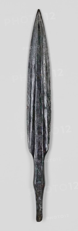 Javelin head, unknown Near Eastern workshop, weapon; flat spear head with an elongated lanceolate blade with a high rib of rectangular cross-section, transitioning at the base into a short, rectangular socket, which transitions into an equally short, tapering, lath-like spike of rectangular cross-section. Luristan bronzes, militaria, ranged weapon, material culture, Eastern Gaulish Scenography. 9th–6th century BC end of the 2nd millennium–first half of the 1st millennium BC; Iron Period. Near East; Luristan. Height 15.3 cm, width 1.8 cm, depth 0.7 cm., An elongated metal object with a pointed tip and a tapered handle, showing an overall dark metallic color with areas of greenish and brownish patina, a central raised ridge running along its length, a textured and slightly irregular surface, and a narrow rounded end opposite the tip, positioned against a pale gray background.