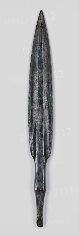 Javelin head, unknown Near Eastern workshop; flat spear head with an elongated lanceolate blade with a high rib of rectangular cross-section, transitioning at the base into a short rectangular socket, which transitions into an equally short, tapering lath-like spike of rectangular cross-section. Material: bronze Luristan bronzes?. Function: weapon, ranged weapon/militaria. Style/period: Iron Age, 9th–6th century BC end of 2nd millennium–first half of 1st millennium BC. Place of origin/find: Luristan, Near East. Dimensions: height 15.3 cm; width 1.8 cm; depth 0.7 cm., A long, narrow metal object with a pointed tip and a tapered base, shown vertically against a light gray background; the surface appears rough and uneven with visible ridges and a central raised ridge running along much of its length; the color is predominantly dark gray to black with areas of greenish patina and small patches of brownish discoloration; the lower section narrows into a thinner, slightly flattened shaft bef...