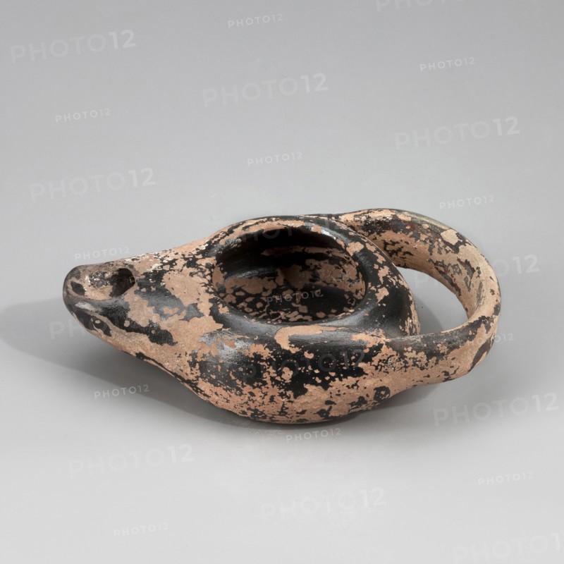 Black-finished lamp, unknown Attic workshop. Lamp made on a wheel. Circular, flattened vessel with curved vertical walls. Flat, circular base. Vertically attached, ribbon-like handle. Arms sloping downward, outward. Large oil hole disc missing. Medium-length burner, slightly upturned. Entire piece coated with black varnish engobe/firnis before firing. Attic clay. 425–400 BC, Myrmekion; Attica origin. Oil lamp; height 2.5 cm, width 5.6 cm, length 9.6 cm., An aged ceramic oil lamp with a rounded body, a raised circular reservoir, a tapered spout, and a curved handle, showing a mottled surface with patches of black and pale terracotta, placed on a smooth light gray background.