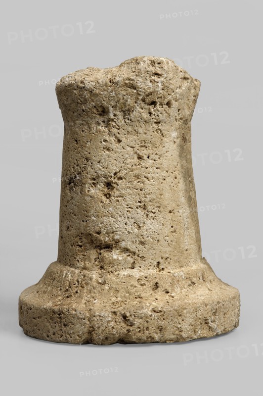 Thymiaterion, missing the upper part of the hearth and approximately half of the base. Thymiaterion with a cylindrical, smooth shaft, slightly tapering towards the top, provided with a high half the height of the shaft profiled base. The base consists of two elements — a vertical lower part and a slightly concave upper part, on which stands the thymiaterion's head, slightly spaced from the edge. Late Hellenistic period, Etruria, varia; height 13.3 cm, width 11 cm., A roughly cylindrical stone object with an irregular, pitted surface sits against a plain light gray background; the stone shows a range of beige, tan, and light brown tones with some darker brown speckles and small cavities, a wider rounded base tapering slightly toward a blunt, uneven top, and visible rough texture and porous holes across its surface.