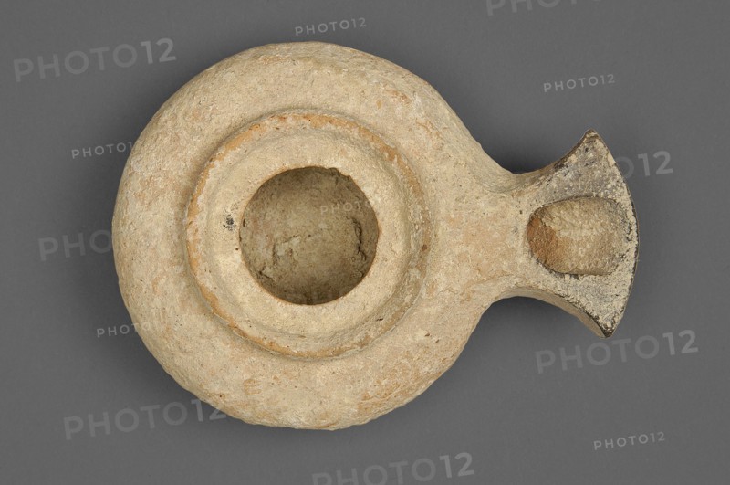 Herodian oil lamp, a small clay lamp with a slightly flattened circular container and a fan-shaped burner. The large round filling opening is surrounded by a convex ring approximately 0.5 cm larger than the opening. Orange clay with light cream engobe. 1st century, Palestine, oil lamp., A round, shallow vessel with a central circular basin and an extended spout-like projection, made of rough, worn material showing a sandy beige and light tan surface with small patches of darker brown and faint orange tones; the rim around the basin is slightly raised, and the basin interior is textured and similar in color to the exterior; the spout area has a darker, weathered edge with mottled gray and brown discoloration; the background is a uniform medium gray.