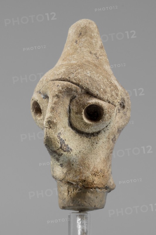 Idol head, unknown Syrian workshop, terracotta, idols, Northwestern Syria. Male head in a conical headdress, rounded at the tip. Flattened, triangular nose, protruding forward. Eyes and ears traces preserved made of glued tablets with a hole drilled in the center. A narrow strip of clay is glued above the forehead. 1st half of the 2nd millennium BC, Orontes Region. Figurine, height 4.5 cm, width 2.5 cm., A close-up of a small sculpted head made of a rough, beige-brown material with weathered texture and irregular surface spots; the head has a pointed top, deep rounded eye sockets set into a pronounced brow ridge, and a long nose extending down toward a small mouth area; the surface shows darker brown and grayish discolorations and patches of lighter beige; the object is mounted on a clear stand against a plain gray background.