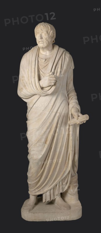 Depiction in the Pseudo-Seneca type, identified as a portrait of Aristophanes or Hesiod; maker, pseudo-Seneca, male portrait in Roman sculpture; togate figure; end of the 1st century BC previously dated 4th quarter of the 1st century BC; Latium; sculpture; weight 610 kg; height 183 cm; width 70 cm; depth 55 cm; base width 62.5 cm; base depth 47 cm., A marble statue of a robed standing man against a dark background, with light beige to off-white stone showing subtle veins and darker weathered spots; the figure has short textured hair and a beard, a solemn facial expression with closed or lightly-open eyes, and a draped cloak wrapped over one shoulder and across the chest with flowing folds and layered fabric; one hand rests near the chest holding part of the drapery while the other extends slightly forward holding a cylindrical object; the lower robes form deep vertical folds that fall to the base, where the statue stands on a rectangular plinth with visible feet and sandals.