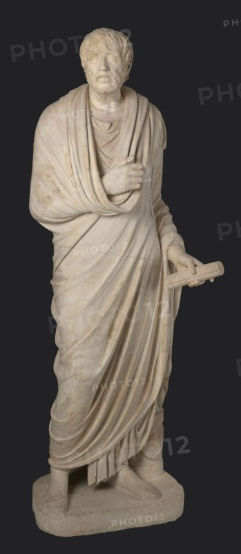 Depiction in the Pseudo-Seneca type, currently identified as a portrait of Aristophanes or Hesiod, pseudo-Seneca, male portrait, Roman sculpture in a toga, end of the 1st century BC previously dated 4th quarter of the 1st century BC, Latium, sculpture; weight 610 kg; height 183 cm; width 70 cm; depth 55 cm; base width 62.5 cm; base depth 47 cm., A full-length pale beige marble statue of a standing robed male figure against a dark gray background, showing a bearded head with short hair and a contemplative facial expression, a draped cloak wrapped over one shoulder and across the chest with carved folds, one hand holding the edge of the cloak near the chest and the other hand extended slightly forward holding a cylindrical scroll, the long garment falling in deep vertical folds over the legs to reveal sandal-clad feet on a rectangular base, with subtle discoloration and veining in the stone.