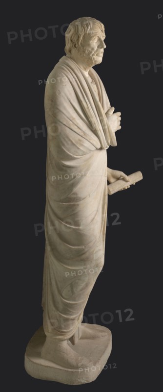 Depiction in the Pseudo-Seneca type, currently identified as a portrait of Aristophanes or Hesiod; creator, pseudo-Seneca, male portrait, Roman sculpture; marble; toga; end of the 1st century BC previously dated to 4th quarter of the 1st century BC; Latium; sculpture; weight 610 kg; height 183 cm; width 70 cm; depth 55 cm; base width 62.5 cm; base depth 47 cm., A full-length pale stone statue of a robed standing man shown in profile against a dark background, with a textured face and short wavy hair, a beard, and a contemplative expression; he is draped in layered flowing garments with visible folds and creases, one arm crossed near his chest and the other extended holding a rolled object; he stands on a rectangular base wearing footwear that is partly visible; the visible colors are pale off-white and beige of the stone, with a dark gray background.
