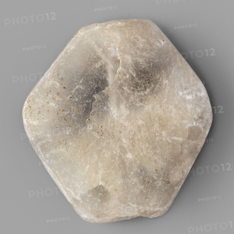 Rhomboid bead, unknown Northern Mesopotamian workshop; rhomboid bead with smooth surfaces, two corners cut off at the ends of the axis, with a drilled hole for stringing. 1st half of the 3rd millennium BC; Early Jazira III period Early Dynastic III; ca. 2600–ca. 2350 BC. Northern Mesopotamia Khabur basin; Tell Rad Shakra northeastern Syria. Found in children's graves. Height 2.2 cm; width 2.08 cm; depth 0.8 cm., A roughly hexagonal stone with rounded edges sits against a flat background; the stone shows a range of muted colors including off-white, beige, gray, and hints of brown, with areas of translucent milky white and darker gray patches, subtle speckles and streaks, and a slightly worn, uneven surface texture.