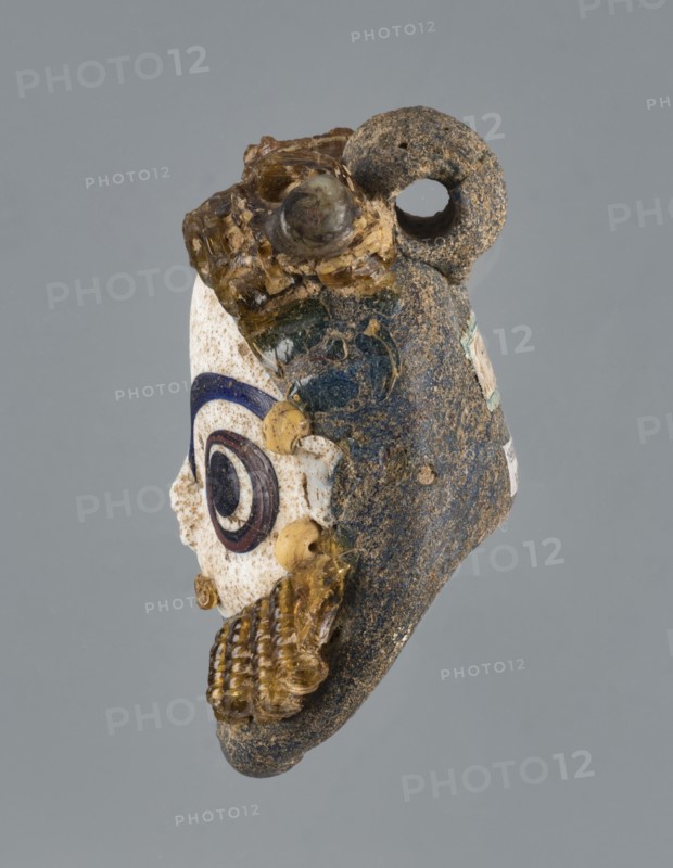 Apotropaic pendant in the shape of a male head, bearded head pendant, pendant in the form of a bearded man's head. 4th–3rd century BC, Carthage Tunisia. Technique on core. Jewelry amulet. Height 8 cm, width 6.4 cm, depth 4 cm., A worn object resembling a small sculpted head with a textured dark gray and brown surface, a loop at the top, and attached translucent yellowish glass elements; one side shows a white area with concentric circular decorations in dark blue and dark brown, and small round yellowish beads; the object displays a mix of colors including gray, brown, white, dark blue, dark brown, and yellowish amber, with rough and smooth textures and some glossy glassy parts.