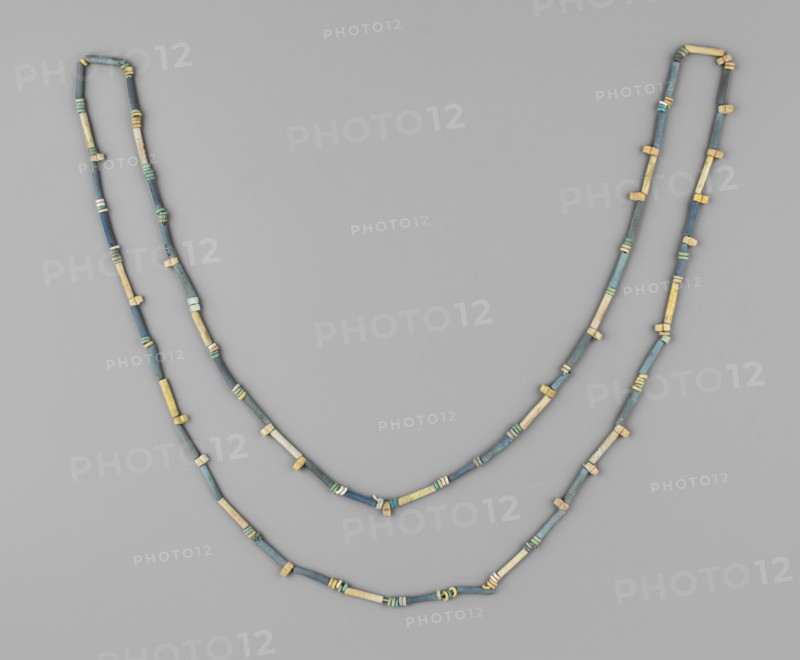 Necklace, disc-shaped and tubular beads; necklace consisting of 52 tubular beads, 118 flat beads and 21 double beads with 2 holes. Colors green and blue, individual beads yellowish. 664–332 BC; Late Period, Egypt. Jewelry; height 113 cm., A pair of beaded necklaces arranged in a loose double loop on a flat background, each necklace made of a sequence of small cylindrical and short tubular beads with occasional slightly larger spacer beads; the beads show a mix of muted blue, teal, green, beige, and pale yellow tones, with some beads appearing weathered or patinated, and the background is a smooth light gray.