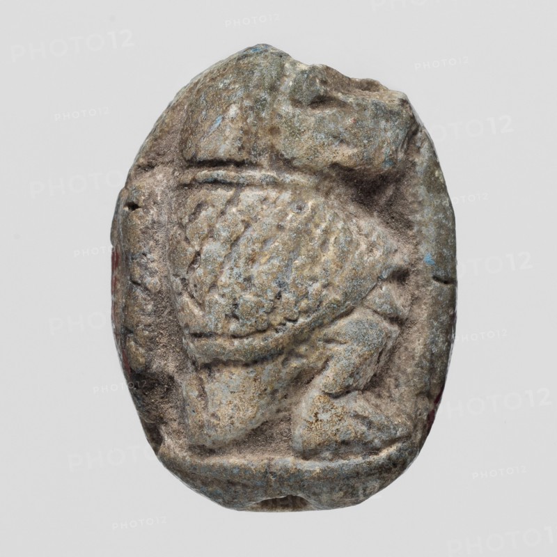 Scaraboid with a monkey. The upper surface of the scaraboid is decorated with a convex image of a seated baboon facing right, with the fur on its back marked with crisscrossing lines. On the base is depicted a lying sphinx lion? wearing a feathered crown, above it a solar disk. In front of the sphinx is the figure of a seated goddess Maat?, above the sphinx's back is visible a lying feather of Maat and possibly another symbol damaged. c. 1550–c. 1186 BC; New Kingdom, 18th–19th Dynasty, Egypt. Amulet, scaraboid; height 2.3 cm, width 1.7 cm., A small worn carved object with an oval outline showing a raised textured central area and surrounding indented grooves, exhibiting a rough surface with pitted and eroded details; the object displays a palette of muted colors including gray, brown, tan, and hints of bluish-green with tiny spots of reddish tone along the edges, and the background is a uniform light gray.