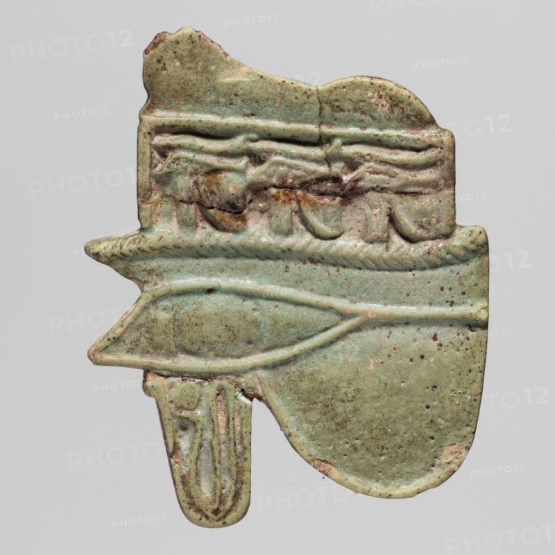 Amulet - Udzhat eye with a lioness;, Udzhat eyes; udjat Eye of Horus and lioness/uraeus iconography; protective amulet used as grave equipment; ca. 945–664 BC, Third Intermediate Period, 22nd–25th Dynasty, Egypt; height 3.6 cm, width 3.8 cm, A greenish, weathered amulet fragment with a matte, speckled surface showing shades of pale green, turquoise, brown, and tan, shaped with a horizontal central eye-like raised motif and a rounded lower lobe; the top edge is irregular with carved relief details including a braided band and draped forms, while the lower portion shows a narrow hanging element with vertical elongated ridges and a chipped edge, and small dark spots and worn areas scattered across the piece.