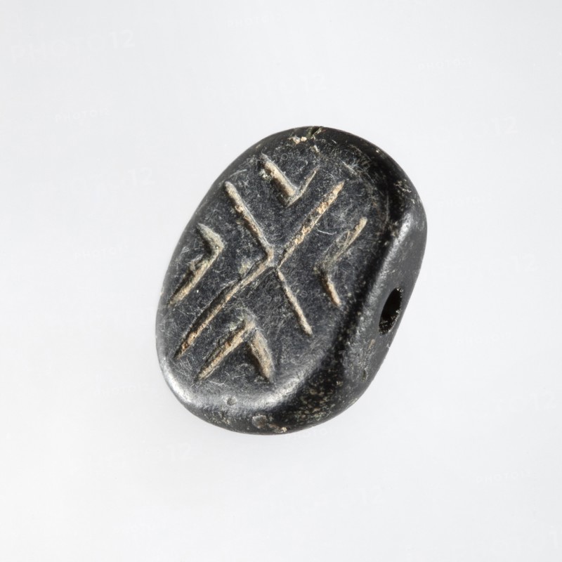 Stamp seal with geometric ornament. A prism-shaped stamp seal with rounded edges and an oval, nearly rectangular base. A hole is drilled through the longer diagonal side walls transverse to the longitudinal axis of the stamp. On a flat base is carved a shallow geometric ornament consisting of regularly spaced, slightly open hooks: two large hooks with their tips touching in the center and four small hooks in triangular fields between the arms of the large hooks. Halaf culture, c. 6th–5th millennium BC. Northern Mesopotamia Iraq, Syria, Turkey. Seal. Height 0.8 cm; width 1.5 cm; depth 1.7 cm; hole diameter 0.2 cm., A small oval object with a smooth, worn surface in dark gray to black tones, showing lighter gray scratches or incisions forming a symmetrical pattern, with a visible rounded edge and a small drilled hole through one side, photographed against a pale, light gray background.